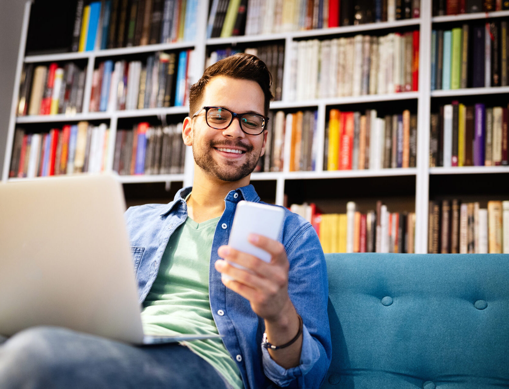 Young man with glasses smiing while looking at his phone at the library