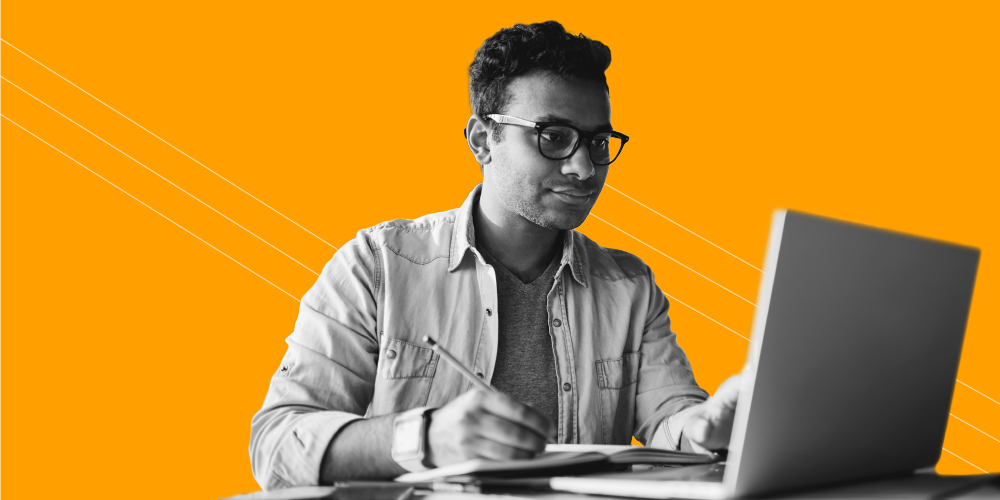 Man with glasses writing with a pencil while working on a laptop