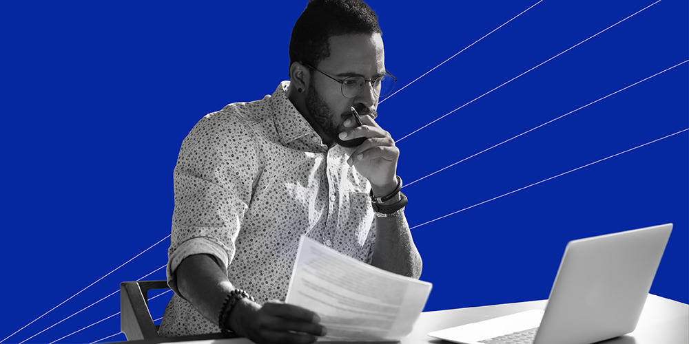man with glasses holding a piece of paper while working on his laptop