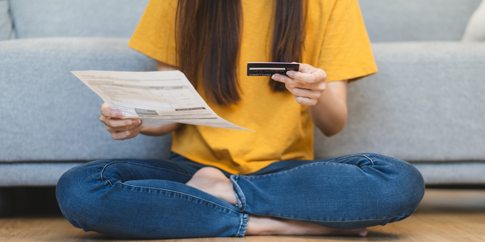 Woman sitting cross legged in front of a couch holding a credit card in one hand and a statement in the other