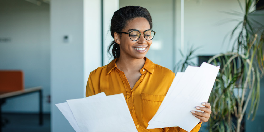 Woman in yellow shirt and glasses smiling while reading a piece of paper